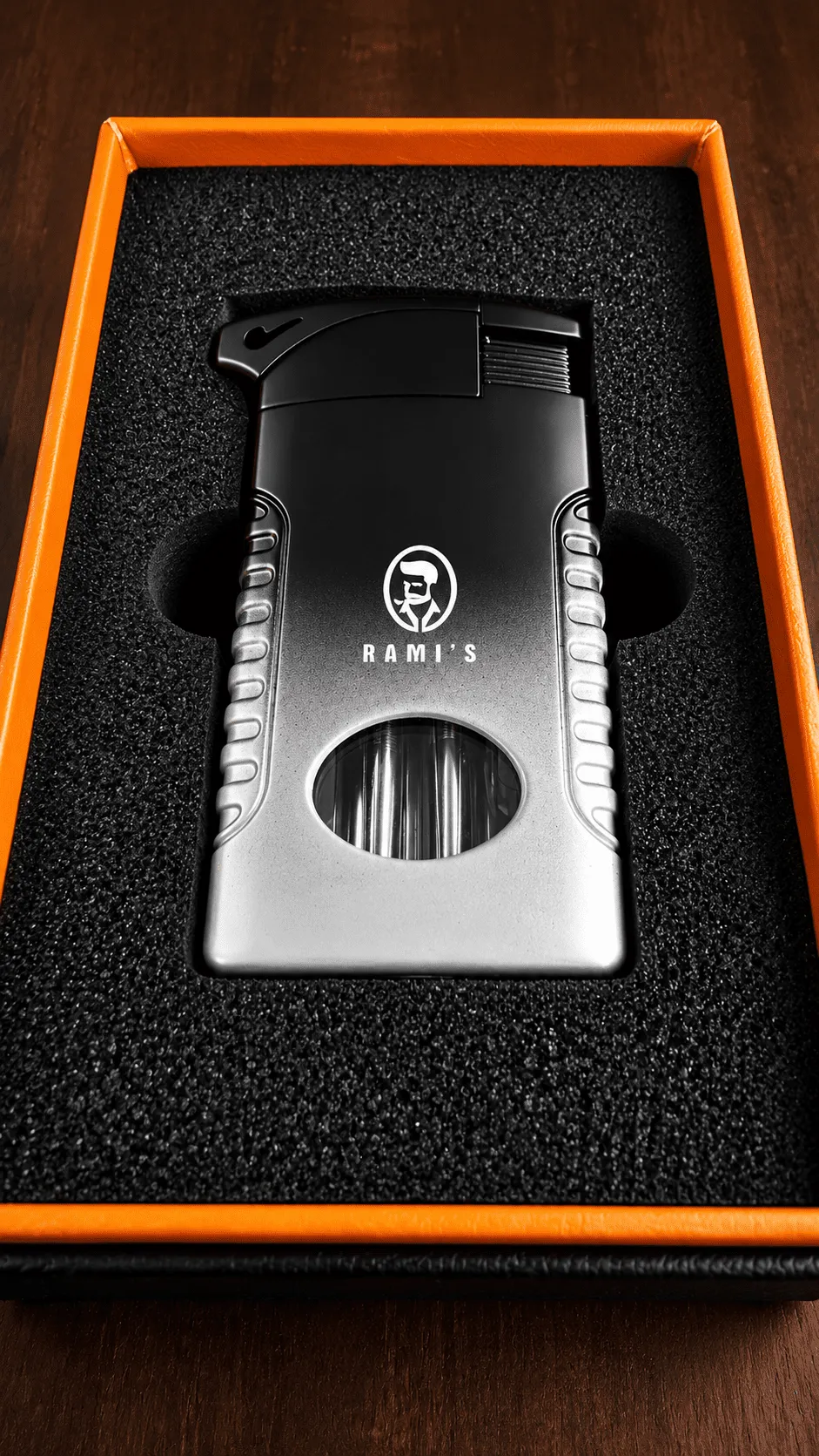 A "Rami's" Pipeliner cigar lighter featuring a sophisticated black-to-silver gradient finish, displayed in a black presentation box with an orange border.