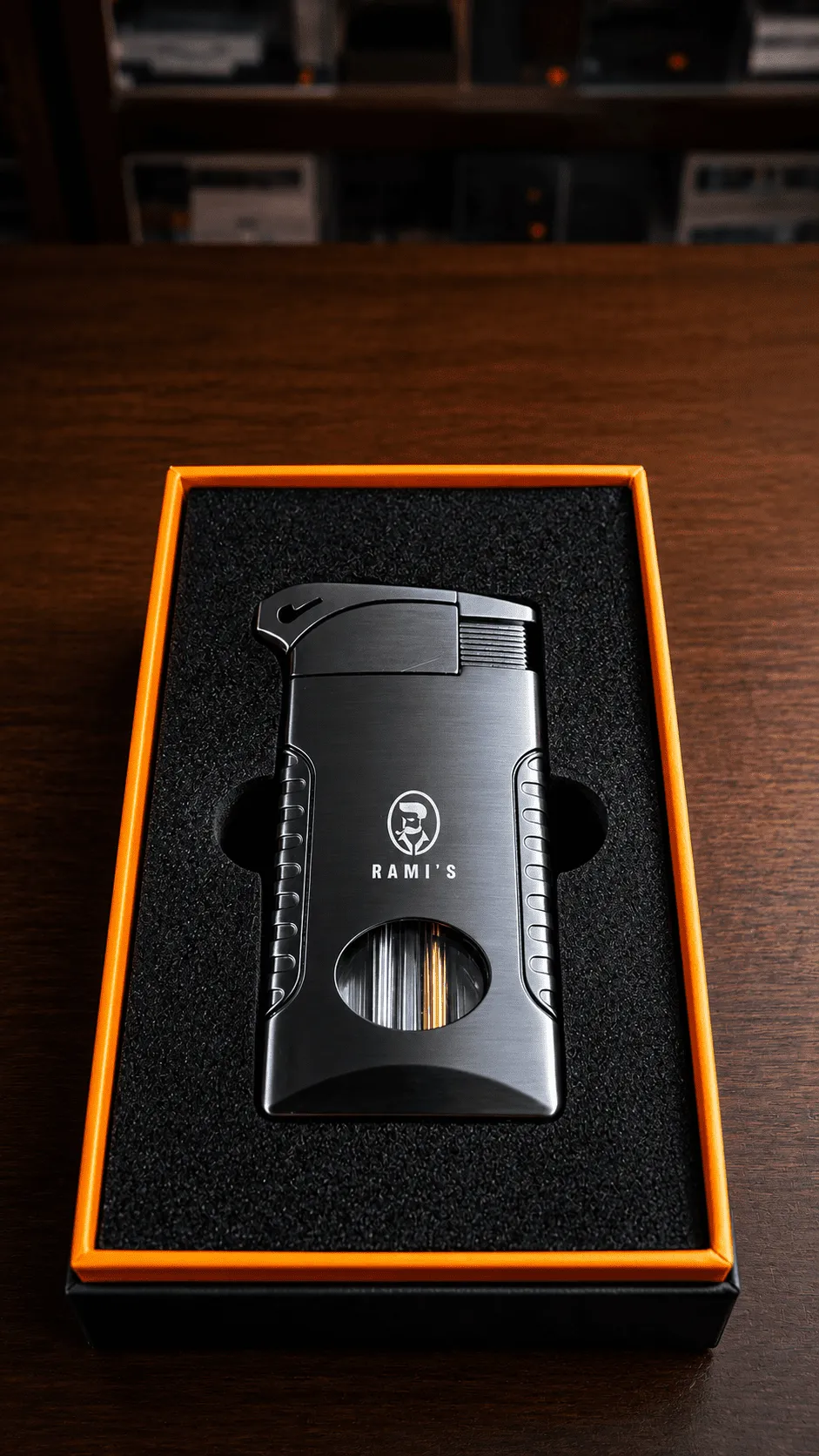 A "Rami's" Pipeliner cigar lighter with a sophisticated, brushed gunmetal finish, presented in a black presentation box with a vibrant orange border