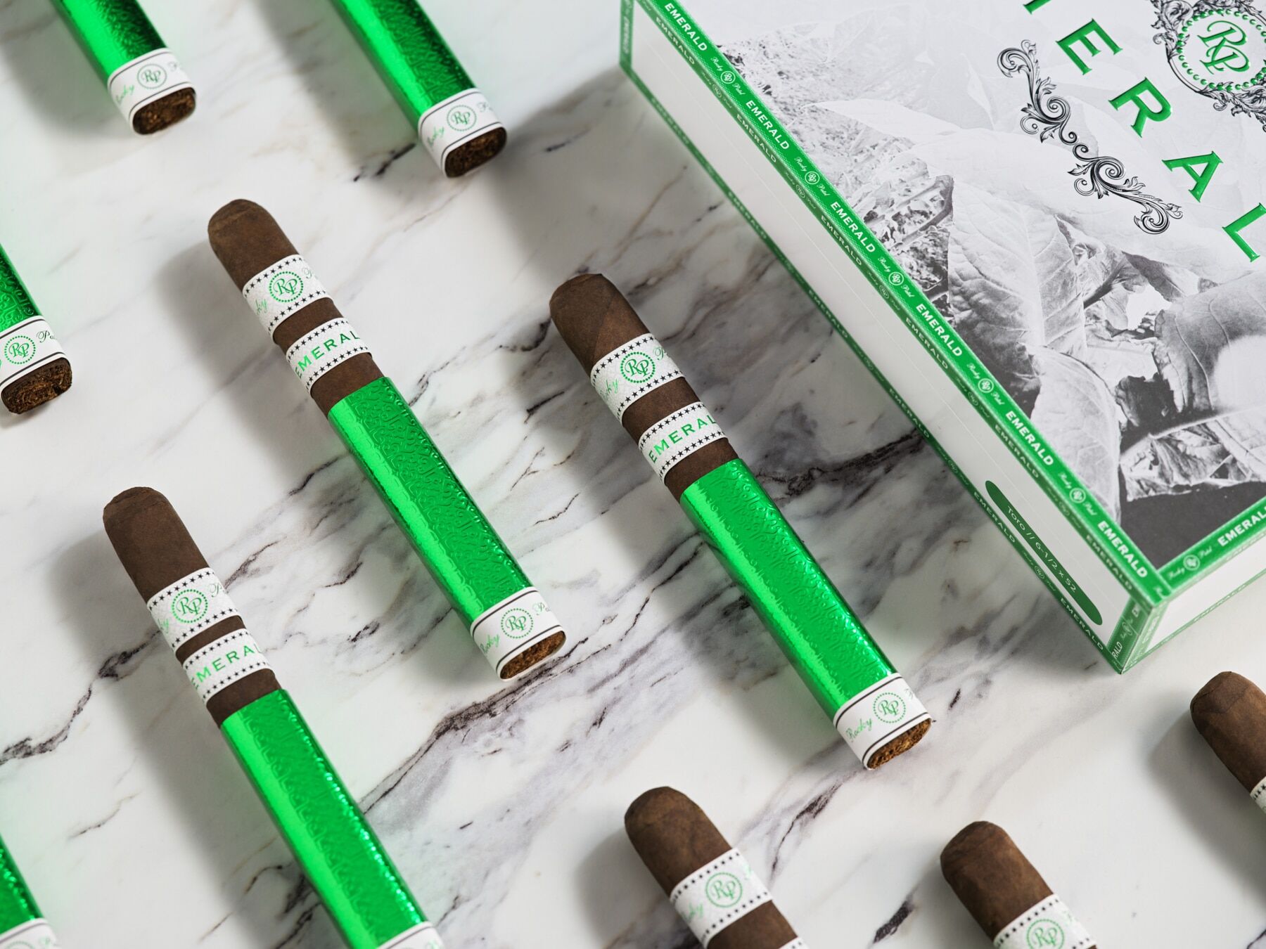 The file Rocky-patel-emerald-cigars-categories.jpg displays multiple box-pressed Rocky Patel Emerald premium cigars laid out on a white marble surface next to a white and green retail display box, showcasing the vibrant green foil sleeve wrapping the lower half of each cigar and the detailed white, green, and gold embossed bands.
