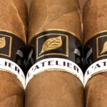 Five L’Atelier L56 cigars with dark, box-pressed wrappers and gold-and-black bands, neatly aligned on a rustic wooden surface.