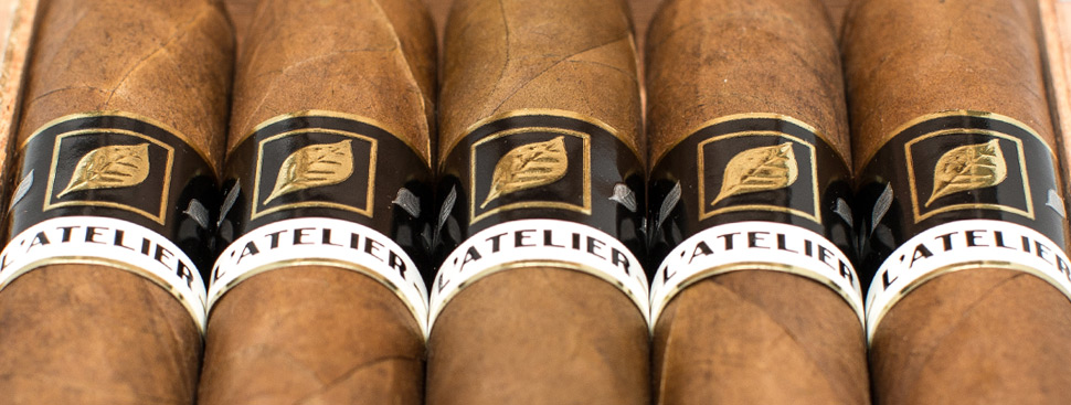 Five L’Atelier L56 cigars with dark, box-pressed wrappers and gold-and-black bands, neatly aligned on a rustic wooden surface.