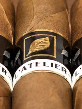 Five L’Atelier L56 cigars with dark, box-pressed wrappers and gold-and-black bands, neatly aligned on a rustic wooden surface.
