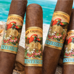 Four San Cristobal Quintessence cigars with dark, oily wrappers and ornate bands resting on a sandy beach with gentle ocean waves
