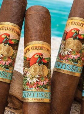 Four San Cristobal Quintessence cigars with dark, oily wrappers and ornate bands resting on a sandy beach with gentle ocean waves