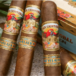 Three San Cristobal Revelation cigars with medium-brown wrappers and ornate bands resting diagonally on a closed wooden cigar box."