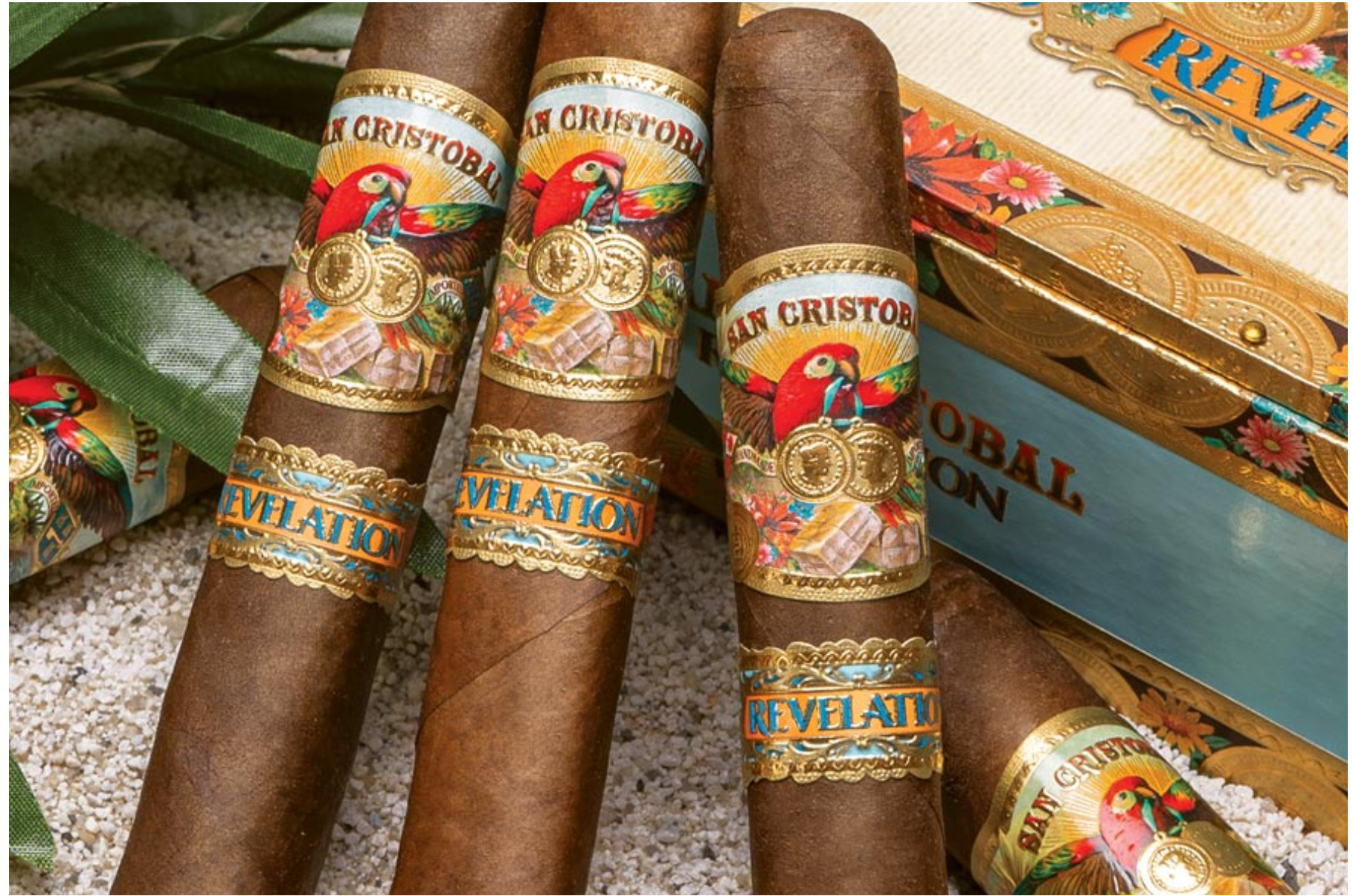 Three San Cristobal Revelation cigars with medium-brown wrappers and ornate bands resting diagonally on a closed wooden cigar box."