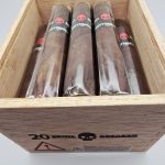 Open box of Tatuaje Surrogates cigars with dark wrappers and bold modern bands arranged in a rustic wooden box.