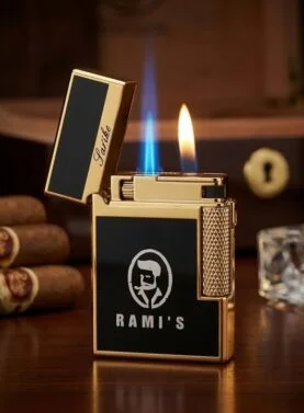 A luxury gold and black Rami's dual-flame lighter featuring a blue torch flame and a soft yellow flame, displayed with premium cigars and a humidor in the background.