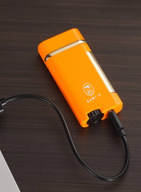 Orange Rami’s dual flame lighter connected to a USB cable, showing rechargeable design and sleek compact body on a desk.