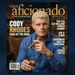 A photograph of the April 2026 issue of Cigar Aficionado magazine featuring Cody Rhodes. He has platinum blonde hair, neck tattoos, and is holding a lit cigar and a glass of whiskey. Headlines include "CODY RHODES KING OF THE RING." The background is a dark blue textured gradient.
