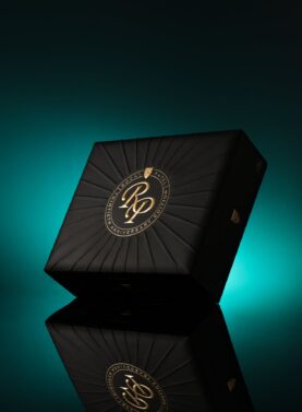 The file Rocky-patel-30th-anniversary-closed-box.jpg displays a closed, premium black wooden retail box of Rocky Patel Thirtieth Anniversary cigars, featuring a textured, sunburst-patterned lid with a central circular gold-embossed "RP Thirtieth Anniversary Edition" logo, standing upright on a reflective black surface against a teal gradient backdrop.