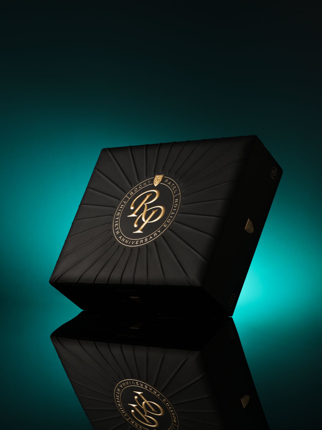 The file Rocky-patel-30th-anniversary-closed-box.jpg displays a closed, premium black wooden retail box of Rocky Patel Thirtieth Anniversary cigars, featuring a textured, sunburst-patterned lid with a central circular gold-embossed "RP Thirtieth Anniversary Edition" logo, standing upright on a reflective black surface against a teal gradient backdrop.