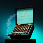 The file Rocky-patel-30th-anniversary-open-box-presentation.jpg displays an open black premium wooden retail box of Rocky Patel Thirtieth Anniversary cigars propped upright, showcasing a ribbed light teal inner lid with a gold-bordered "Rocky Patel Thirtieth Anniversary" logo plate, a row of box-pressed cigars inside, and a single lit cigar resting across the front left emitting a plume of blue-tinted smoke against a teal gradient backdrop