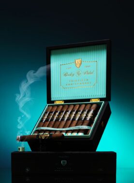 The file Rocky-patel-30th-anniversary-open-box-presentation.jpg displays an open black premium wooden retail box of Rocky Patel Thirtieth Anniversary cigars propped upright, showcasing a ribbed light teal inner lid with a gold-bordered "Rocky Patel Thirtieth Anniversary" logo plate, a row of box-pressed cigars inside, and a single lit cigar resting across the front left emitting a plume of blue-tinted smoke against a teal gradient backdrop