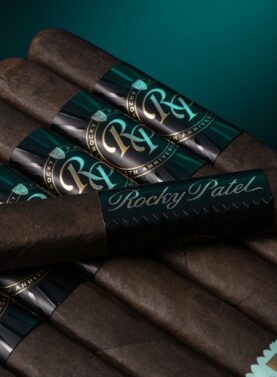 The file Rocky-patel-30th-anniversary-single-cigar.jpg displays a single box-pressed Rocky Patel Thirtieth Anniversary cigar laid horizontally across a stacked bundle of identical cigars on a dark surface, highlighting its rich, dark wrapper, an oversized black band with teal script reading Rocky Patel, and a light teal foot band