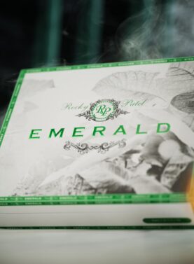 The file Rocky-patel-emerald-closed-box.jpg displays a closed, premium white wooden retail box of Rocky Patel Emerald cigars, featuring a white lid with a black-and-white tobacco leaf pattern, bold metallic green EMERALD branding, a central green RP logo, and ambient smoke swirling around the packaging against a dark backdrop.