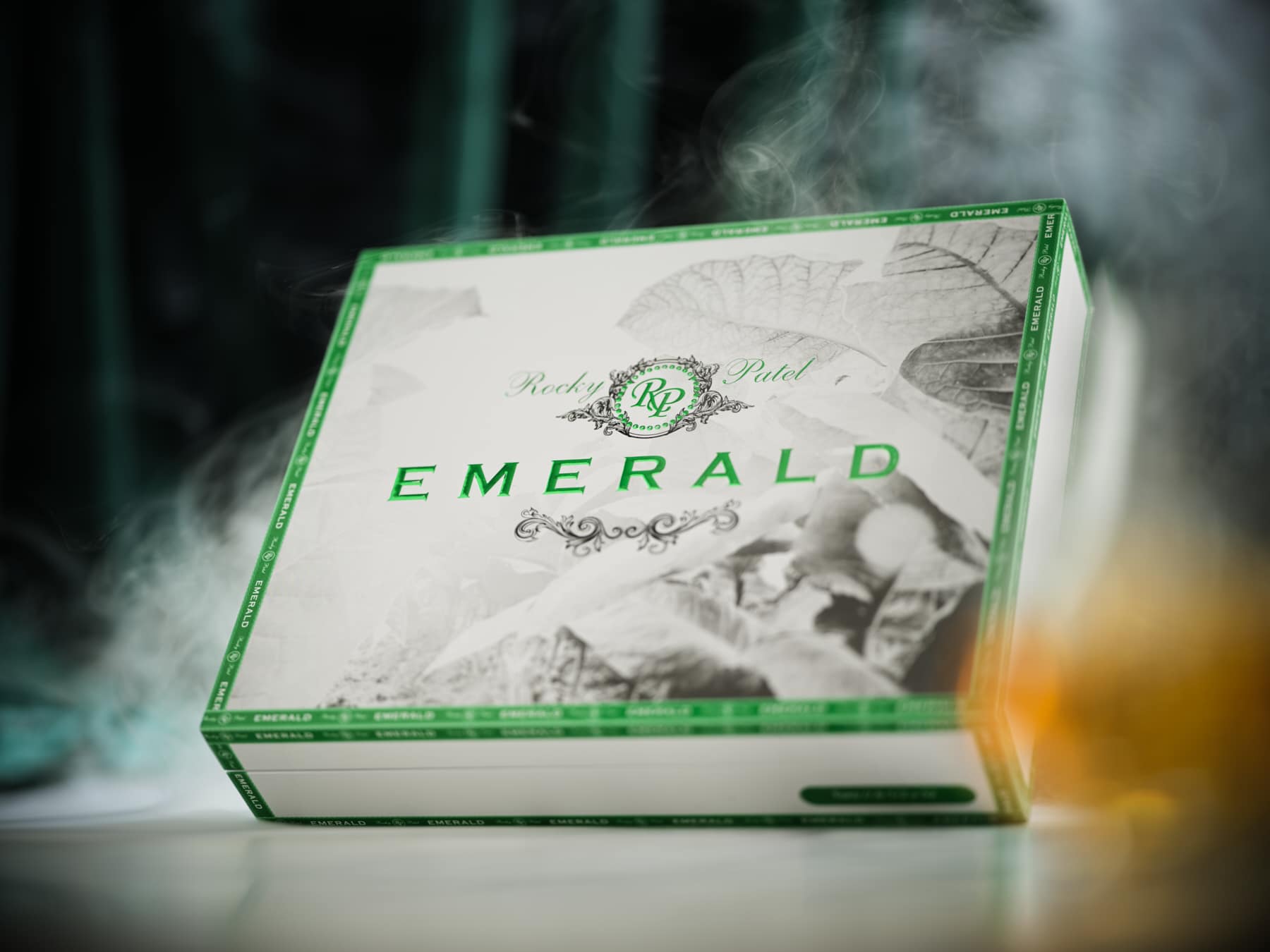 The file Rocky-patel-emerald-closed-box.jpg displays a closed, premium white wooden retail box of Rocky Patel Emerald cigars, featuring a white lid with a black-and-white tobacco leaf pattern, bold metallic green EMERALD branding, a central green RP logo, and ambient smoke swirling around the packaging against a dark backdrop.