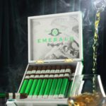 The file Rocky-patel-emerald-open-box-presentation.jpg displays an upright, open white wooden retail box of Rocky Patel Emerald cigars on a white marble surface, showcasing the inner lid design, row of foil-wrapped sticks, a single cigar resting on a stand to the left, and amber liquid pouring into a lowball glass on the right amid ambient smoke and a dark green backdrop.