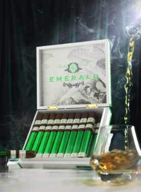 The file Rocky-patel-emerald-open-box-presentation.jpg displays an upright, open white wooden retail box of Rocky Patel Emerald cigars on a white marble surface, showcasing the inner lid design, row of foil-wrapped sticks, a single cigar resting on a stand to the left, and amber liquid pouring into a lowball glass on the right amid ambient smoke and a dark green backdrop.