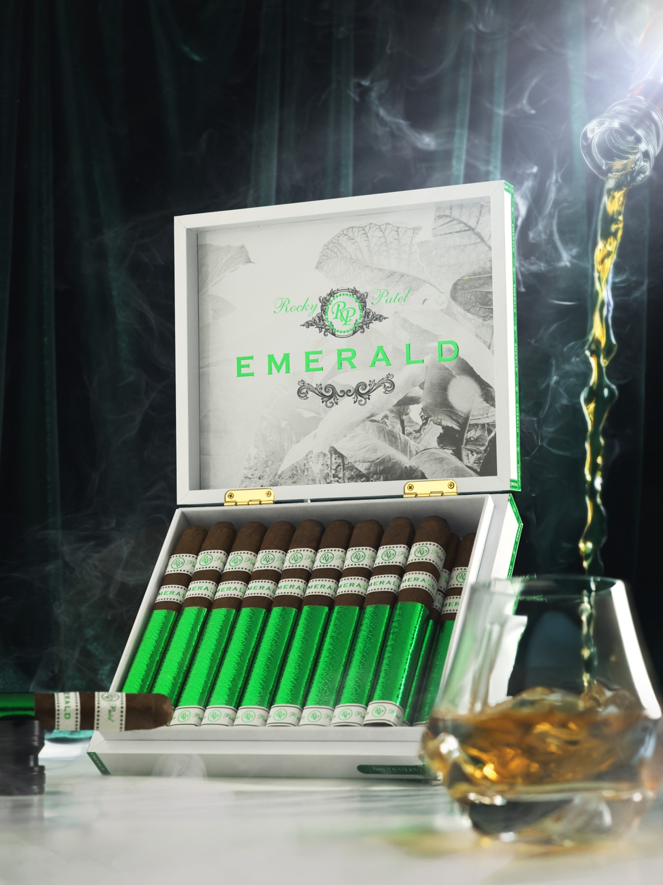 The file Rocky-patel-emerald-open-box-presentation.jpg displays an upright, open white wooden retail box of Rocky Patel Emerald cigars on a white marble surface, showcasing the inner lid design, row of foil-wrapped sticks, a single cigar resting on a stand to the left, and amber liquid pouring into a lowball glass on the right amid ambient smoke and a dark green backdrop.