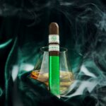 The file Rocky-patel-emerald-single-cigar.jpg displays a single, upright box-pressed Rocky Patel Emerald premium cigar propped against a lowball glass containing amber bourbon, set against a draped dark green velvet backdrop with thick swirls of white smoke