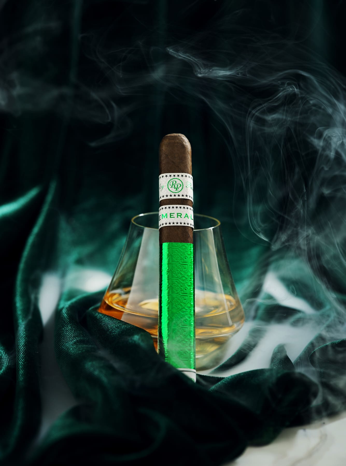 The file Rocky-patel-emerald-single-cigar.jpg displays a single, upright box-pressed Rocky Patel Emerald premium cigar propped against a lowball glass containing amber bourbon, set against a draped dark green velvet backdrop with thick swirls of white smoke
