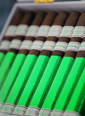 The file Rocky-patel-open-box-presentation- close-shot.jpg displays a close-up, angled view of an open white retail box filled with Rocky Patel Emerald cigars, highlighting the vibrant green foil sleeves on each stick, white and green embossed dual bands, and a soft wisp of ambient smoke drifting across the left side of the frame.