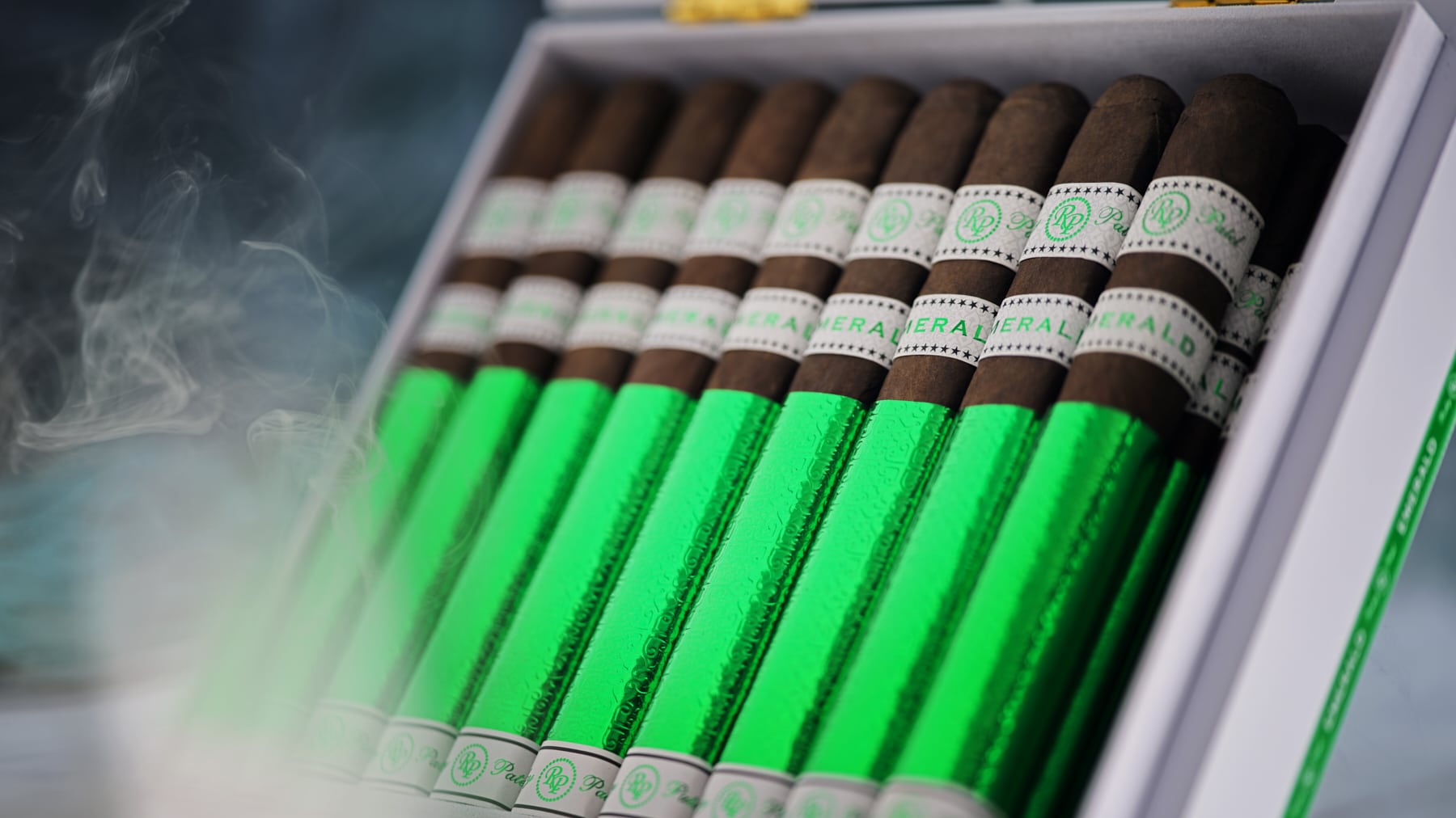 The file Rocky-patel-open-box-presentation- close-shot.jpg displays a close-up, angled view of an open white retail box filled with Rocky Patel Emerald cigars, highlighting the vibrant green foil sleeves on each stick, white and green embossed dual bands, and a soft wisp of ambient smoke drifting across the left side of the frame.