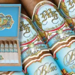 E-commerce promotional banner featuring the new My Father Blue Honduras cigars, showcasing an open display box, detailed views of the light blue bands with Roman numerals MMXXV, and a rustic wooden background
