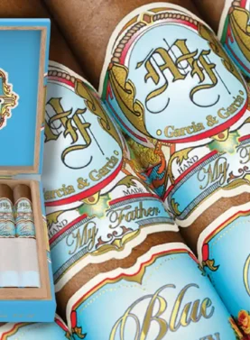 E-commerce promotional banner featuring the new My Father Blue Honduras cigars, showcasing an open display box, detailed views of the light blue bands with Roman numerals MMXXV, and a rustic wooden background