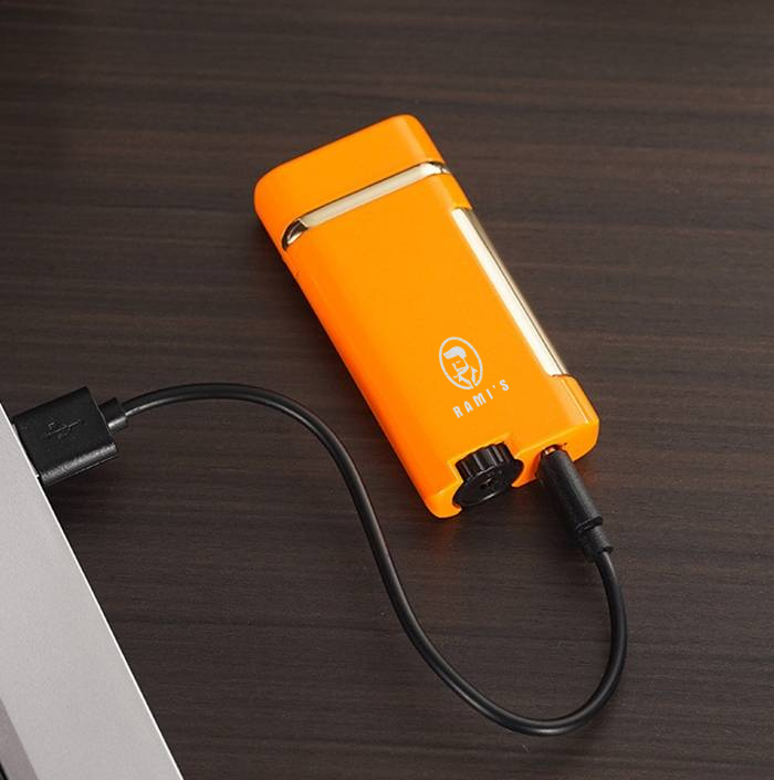 A vibrant orange Rami's Accessories lighter with gold trim, resting on a dark wood surface while connected to a laptop via a USB charging cable.