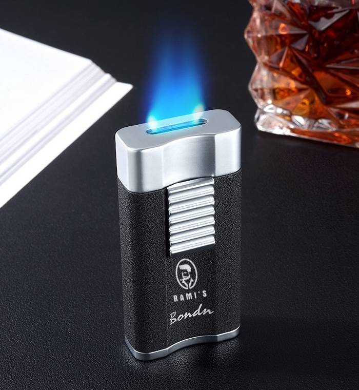 A textured black and silver Rami's Bondu cigar lighter emitting a wide, flat blue waterfall-style torch flame, positioned next to a glass of bourbon.