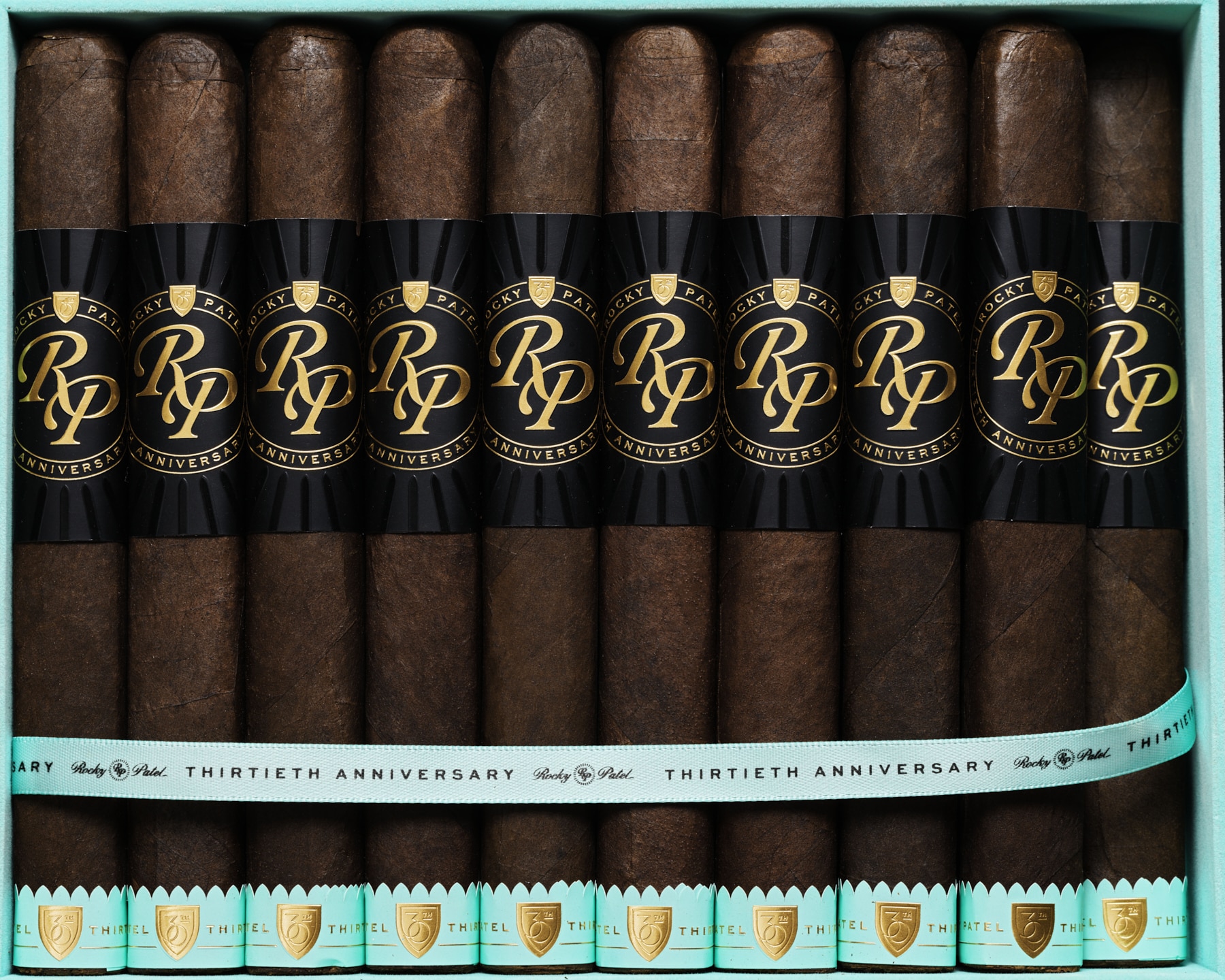 The file rocky-patel-30th-annivesary-category-product.jpg displays a top-down, close-up view of ten premium box-pressed Rocky Patel Thirtieth Anniversary cigars neatly arranged side-by-side inside a light teal retail display box, showcasing their dark oily wrappers, prominent black and gold embossed "RP Anniversary" main bands, a matching light teal ribbon across the center, and teal and gold foot bands.
