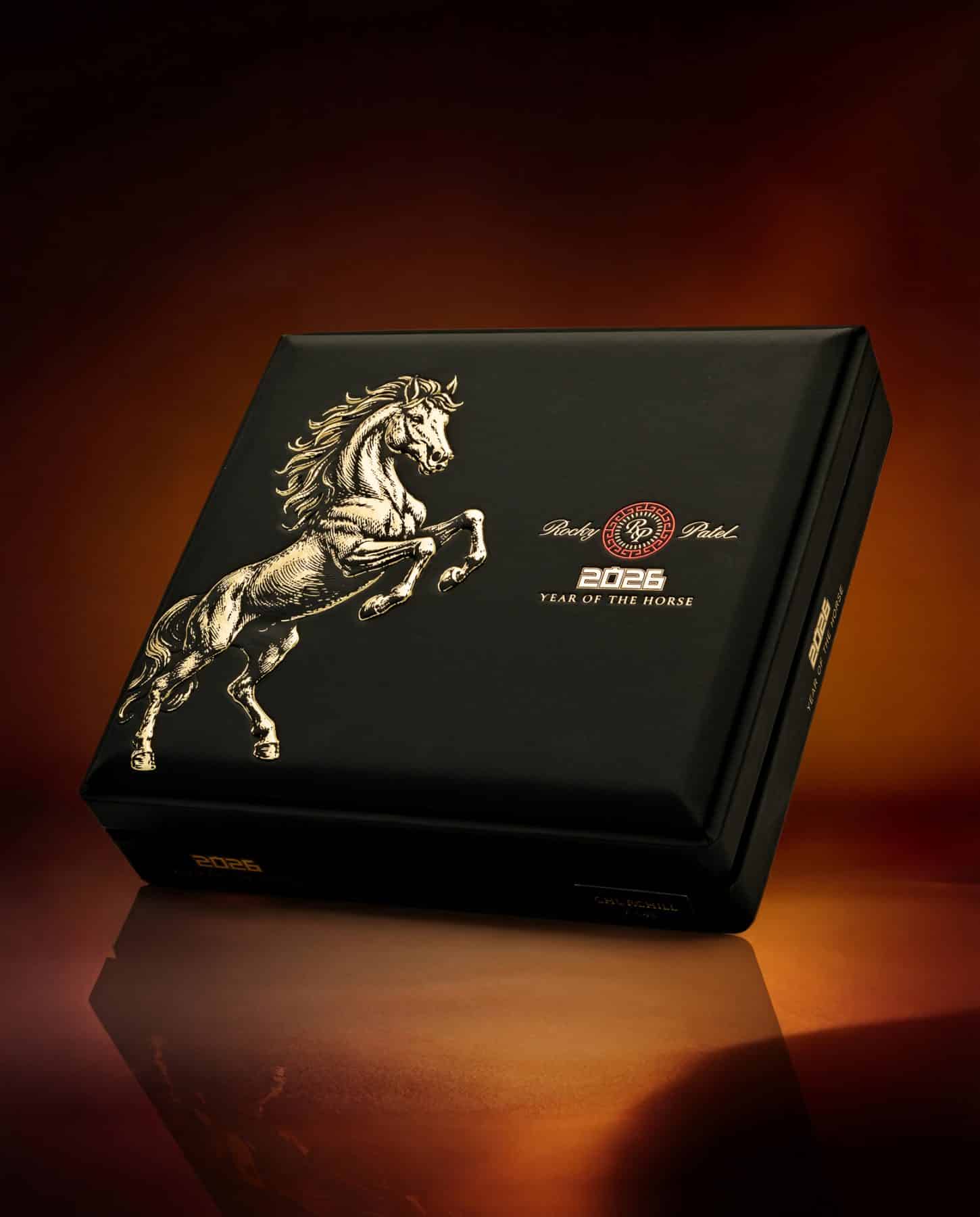 The file rocky-patel-cigar-year-of-the-horse-closed-box.jpg displays a closed, premium matte black wooden retail box of Rocky Patel 2026 Year of the Horse cigars, featuring a detailed gold-foil illustration of a rearing horse on the left side of the lid, a red-and-gold central Rocky Patel logo, and "2026 Year of the Horse" text, standing at an angle on a highly reflective surface against a warm brown gradient background.