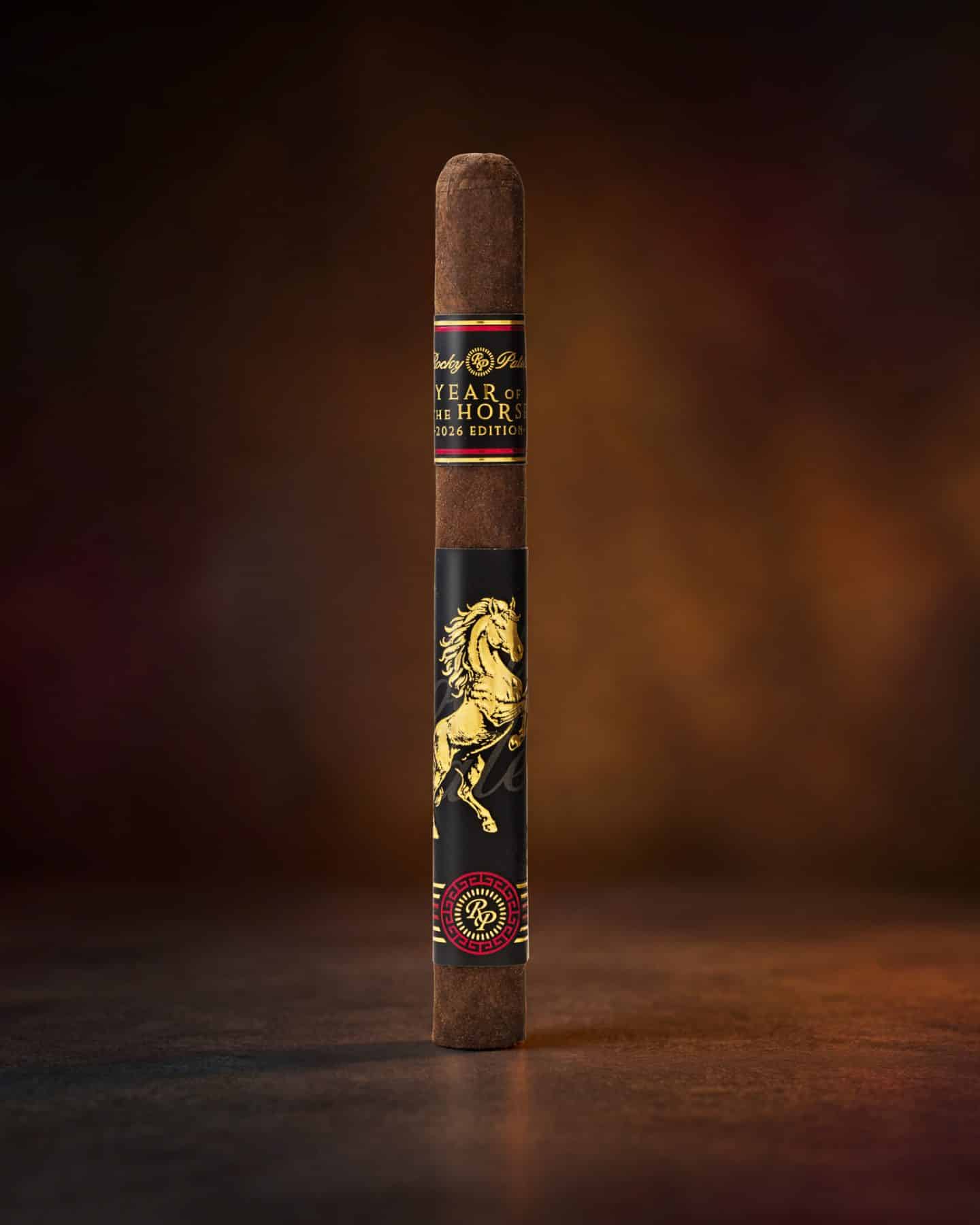 The file rocky-patel-cigar-year-of the horse-single.jpg displays a single, upright Rocky Patel Year of the Horse 2026 Edition premium cigar standing vertically on a textured surface, showcasing its dark wrapper, a top black-and-gold band inscribed with "Year of the Horse 2026 Edition," a large central black sleeve featuring a gold rearing horse illustration, and a detailed red-and-gold foot band against a warm, mottled brown background.