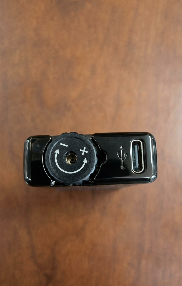 A top-down macro shot of the bottom base of a glossy black Rami's Accessories lighter, showcasing a prominent circular flame adjustment wheel with plus and minus indicators, a central butane refill valve, and a modern USB-C charging port.